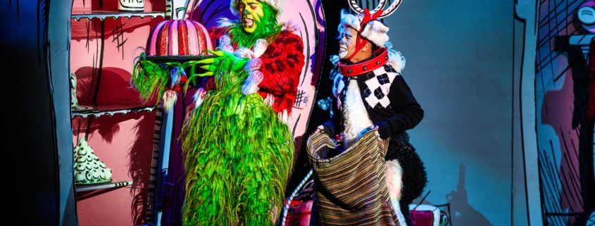 The Grinch, dressed in a green furry costume with a red Santa hat, holds a large pink and white striped cake on a platter. Next to the Grinch is another character dressed as Max the dog, wearing antlers and holding a sack. They are standing in front of colorful cartoon-like scenery from Whoville.