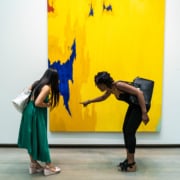 Two people viewing a large abstract painting with a bright yellow background, blue patches, and small red areas in a gallery; one person points while the other leans in to observe.