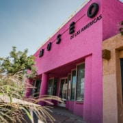 Exterior view of Museo de las Americas, a vibrant pink building with bold black lettering and surrounding greenery under a clear blue sky.