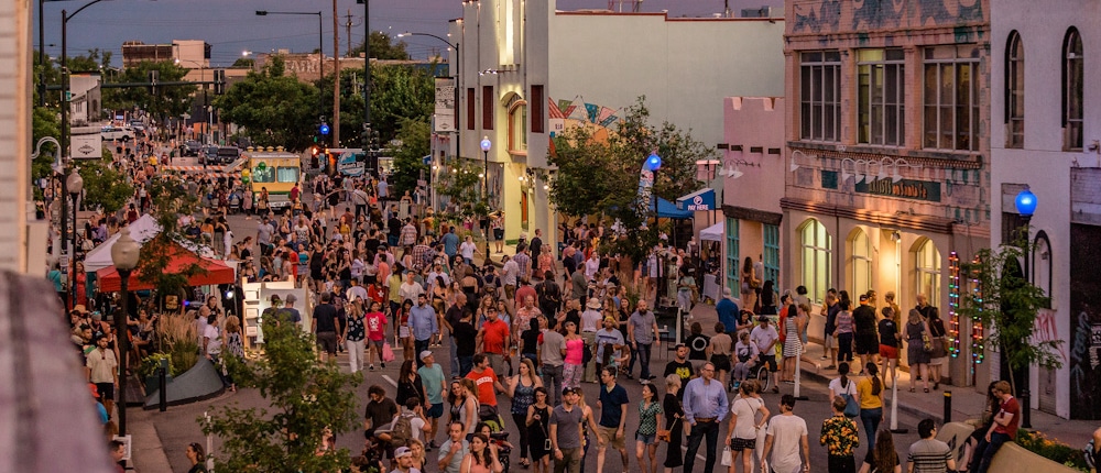 Bustling street festival with crowds walking among colorful buildings, murals, and vendor booths under a purple-pink sky at dawn or dusk.