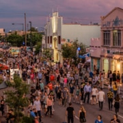 Bustling street festival with crowds walking among colorful buildings, murals, and vendor booths under a purple-pink sky at dawn or dusk.