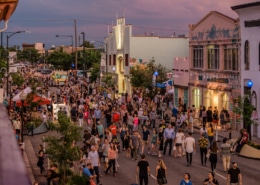 Bustling street festival with crowds walking among colorful buildings, murals, and vendor booths under a purple-pink sky at dawn or dusk.