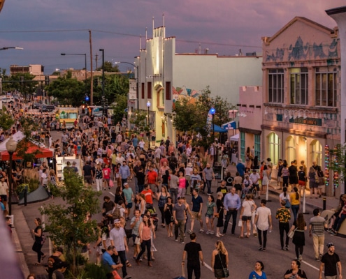 Bustling street festival with crowds walking among colorful buildings, murals, and vendor booths under a purple-pink sky at dawn or dusk.