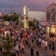 Bustling street festival with crowds walking among colorful buildings, murals, and vendor booths under a purple-pink sky at dawn or dusk.