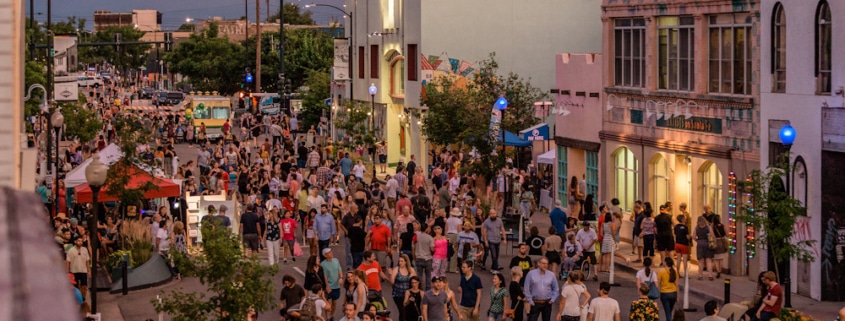 Bustling street festival with crowds walking among colorful buildings, murals, and vendor booths under a purple-pink sky at dawn or dusk.