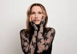 Portrait of a person wearing a black lace outfit, posing with hands touching face against a plain background.
