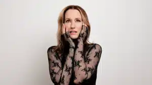 Portrait of a person wearing a black lace outfit, posing with hands touching face against a plain background.