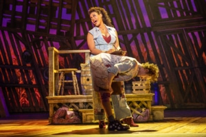 Two performers on stage in a theatrical production of Shucked. One stands and sings while the other bends over in a comedic or dramatic pose, set against a rustic backdrop with wooden structures and props.