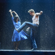 Two dancers performing energetically in the rain on a stage, with dramatic lighting and water splashing around them