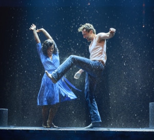 Two dancers performing energetically in the rain on a stage, with dramatic lighting and water splashing around them