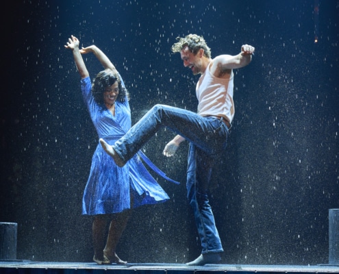 Two dancers performing energetically in the rain on a stage, with dramatic lighting and water splashing around them