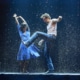 Two dancers performing energetically in the rain on a stage, with dramatic lighting and water splashing around them