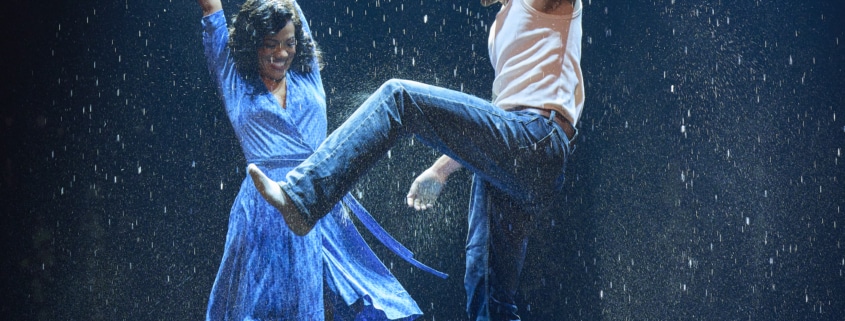 Two dancers performing energetically in the rain on a stage, with dramatic lighting and water splashing around them
