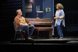 Two people on stage, one sitting at a piano and the other standing, engaged in conversation. The setting includes a dimly lit room with various objects and furniture.