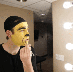 An actor in front of a dressing table painting his face green and black to portray the Grinch