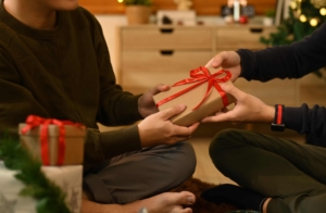Two people sitting on the floor exchanging a wrapped gift with a red ribbon in a cozy, Christmas-themed indoor setting