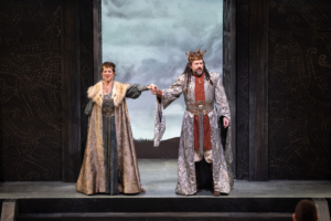 Two actors in elaborate medieval-style costumes stand on a stage holding hands. One wears a long fur-trimmed gown with intricate patterns, and the other is dressed in a regal robe with a crown and ornate belt. The backdrop shows a cloudy sky and distant hills, framed by dark panels with decorative carvings.