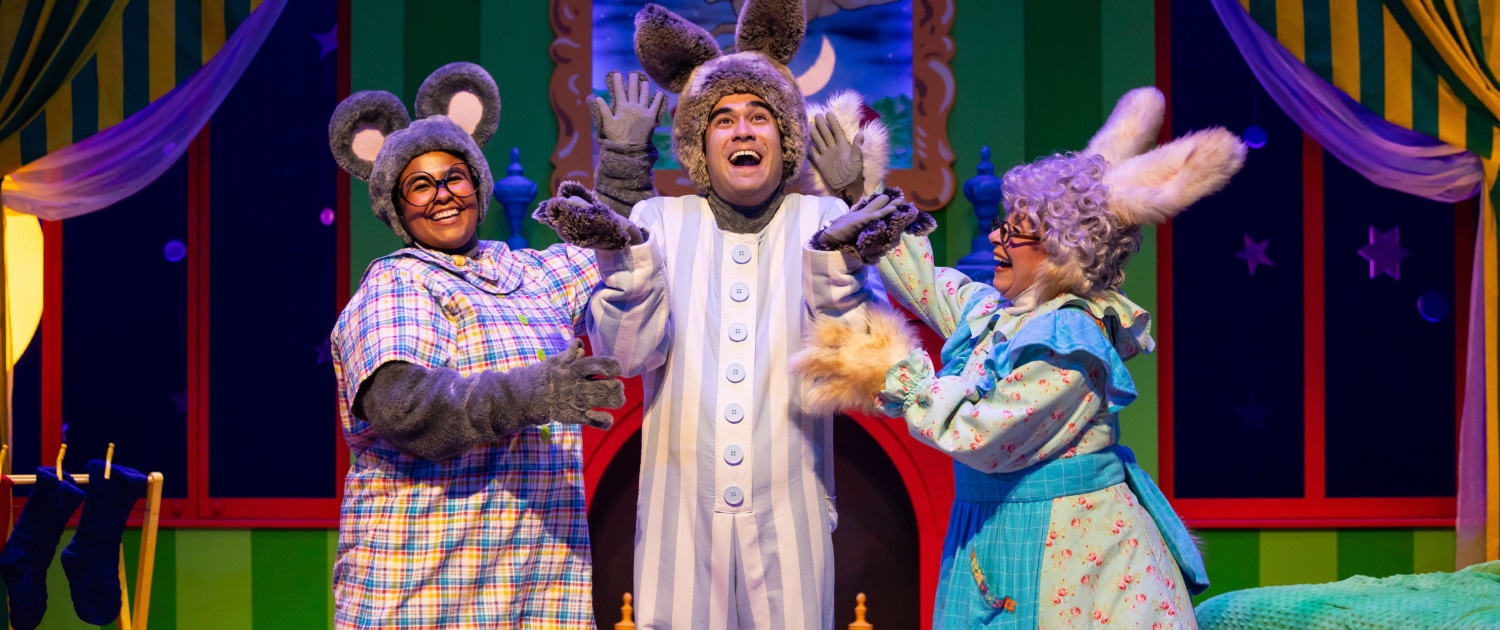 Three performers in animal costumes on a colorful stage set resembling a bedroom from a children’s story. One wears gray mouse ears and a plaid shirt, another has tall bunny ears and striped pajamas, and the third wears a blue floral dress with fluffy cuffs and rabbit ears. The background features green striped walls, yellow curtains, a crescent moon in the window, and a cozy bed with a green blanket.