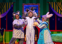 Three performers in animal costumes on a colorful stage set resembling a bedroom from a children’s story. One wears gray mouse ears and a plaid shirt, another has tall bunny ears and striped pajamas, and the third wears a blue floral dress with fluffy cuffs and rabbit ears. The background features green striped walls, yellow curtains, a crescent moon in the window, and a cozy bed with a green blanket.