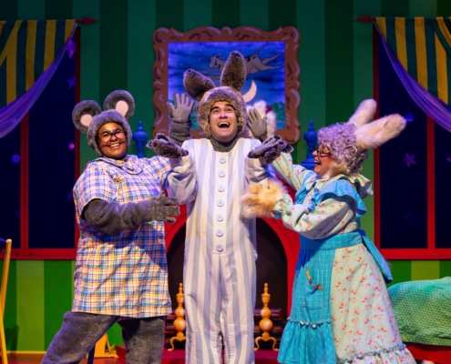 Three performers in animal costumes on a colorful stage set resembling a bedroom from a children’s story. One wears gray mouse ears and a plaid shirt, another has tall bunny ears and striped pajamas, and the third wears a blue floral dress with fluffy cuffs and rabbit ears. The background features green striped walls, yellow curtains, a crescent moon in the window, and a cozy bed with a green blanket.