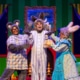 Three performers in animal costumes on a colorful stage set resembling a bedroom from a children’s story. One wears gray mouse ears and a plaid shirt, another has tall bunny ears and striped pajamas, and the third wears a blue floral dress with fluffy cuffs and rabbit ears. The background features green striped walls, yellow curtains, a crescent moon in the window, and a cozy bed with a green blanket.