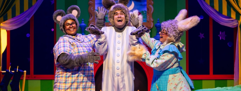 Three performers in animal costumes on a colorful stage set resembling a bedroom from a children’s story. One wears gray mouse ears and a plaid shirt, another has tall bunny ears and striped pajamas, and the third wears a blue floral dress with fluffy cuffs and rabbit ears. The background features green striped walls, yellow curtains, a crescent moon in the window, and a cozy bed with a green blanket.
