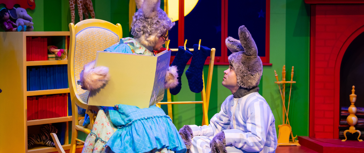 Stage scene from a colorful children’s play featuring two characters dressed as rabbits; one sits in a rocking chair holding an oversized book, while the other sits on the floor in striped pajamas. The background includes a bright green wall, a red fireplace, stockings, and a large yellow moon visible through a window.