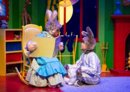 Stage scene from a colorful children’s play featuring two characters dressed as rabbits; one sits in a rocking chair holding an oversized book, while the other sits on the floor in striped pajamas. The background includes a bright green wall, a red fireplace, stockings, and a large yellow moon visible through a window.