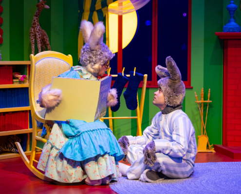 Stage scene from a colorful children’s play featuring two characters dressed as rabbits; one sits in a rocking chair holding an oversized book, while the other sits on the floor in striped pajamas. The background includes a bright green wall, a red fireplace, stockings, and a large yellow moon visible through a window.