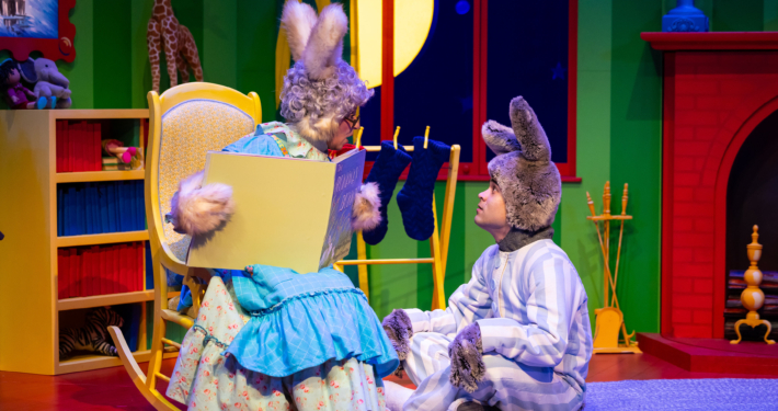 Stage scene from a colorful children’s play featuring two characters dressed as rabbits; one sits in a rocking chair holding an oversized book, while the other sits on the floor in striped pajamas. The background includes a bright green wall, a red fireplace, stockings, and a large yellow moon visible through a window.