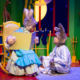 Stage scene from a colorful children’s play featuring two characters dressed as rabbits; one sits in a rocking chair holding an oversized book, while the other sits on the floor in striped pajamas. The background includes a bright green wall, a red fireplace, stockings, and a large yellow moon visible through a window.