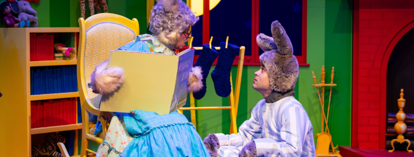 Stage scene from a colorful children’s play featuring two characters dressed as rabbits; one sits in a rocking chair holding an oversized book, while the other sits on the floor in striped pajamas. The background includes a bright green wall, a red fireplace, stockings, and a large yellow moon visible through a window.