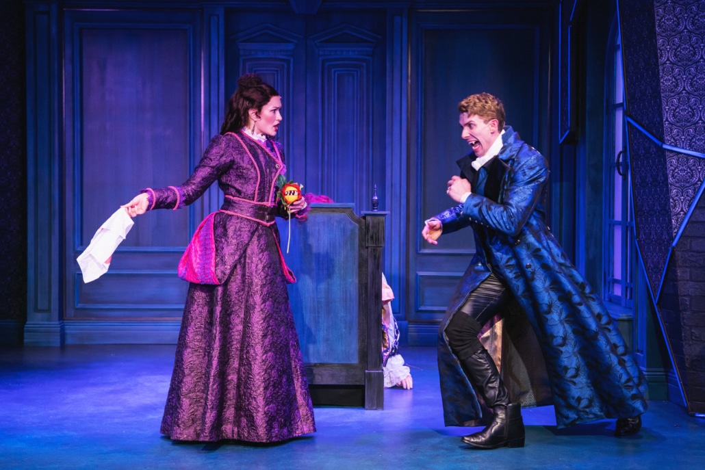 Two actors on stage in an ornate, blue-toned set, wearing elaborate period costumes; one holds a piece of paper while the other gestures dramatically, suggesting a tense or comedic moment in a theatrical performance.
