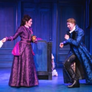 Two actors on stage in an ornate, blue-toned set, wearing elaborate period costumes; one holds a piece of paper while the other gestures dramatically, suggesting a tense or comedic moment in a theatrical performance.