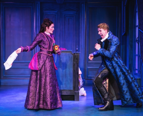 Two actors on stage in an ornate, blue-toned set, wearing elaborate period costumes; one holds a piece of paper while the other gestures dramatically, suggesting a tense or comedic moment in a theatrical performance.