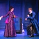Two actors on stage in an ornate, blue-toned set, wearing elaborate period costumes; one holds a piece of paper while the other gestures dramatically, suggesting a tense or comedic moment in a theatrical performance.