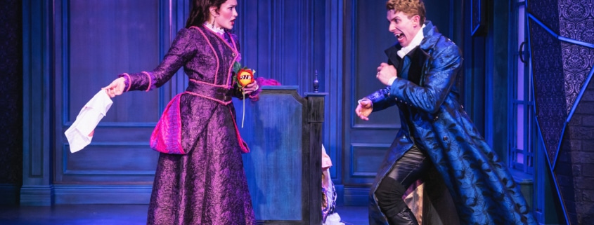 Two actors on stage in an ornate, blue-toned set, wearing elaborate period costumes; one holds a piece of paper while the other gestures dramatically, suggesting a tense or comedic moment in a theatrical performance.