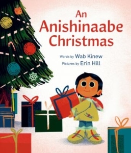 Book cover showing a child holding a red gift box in front of a decorated Christmas tree with colorful presents underneath. Title reads “An Anishinaabe Christmas” with words by Wab Kinew and pictures by Erin Hill.