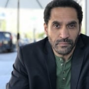 Person sitting at an outdoor café table wearing a dark blazer over a green henley shirt, with a blurred city street and parked car in the background.