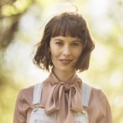Person outdoors wearing a light denim overall layered over a silky blouse with a large bow tie at the neck, with sunlit trees in the background.