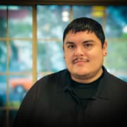 Person indoors wearing a black collared shirt, standing in front of a large window with a blurred outdoor view featuring greenery and a parked orange vehicle.