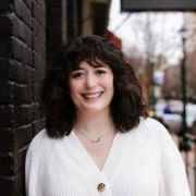 Person standing against a black brick wall wearing a white textured cardigan with a single large button, urban street scene in the background.