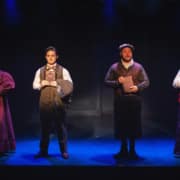 Four actors stand in a row on a dimly lit stage, each holding a book in front of their chest. They are dressed in Victorian-style costumes, including long skirts, vests, and high-collared blouses, against a dark backdrop with subtle paneling. The lighting creates a dramatic, moody atmosphere with blue and purple tones.