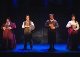 Four actors stand in a row on a dimly lit stage, each holding a book in front of their chest. They are dressed in Victorian-style costumes, including long skirts, vests, and high-collared blouses, against a dark backdrop with subtle paneling. The lighting creates a dramatic, moody atmosphere with blue and purple tones.