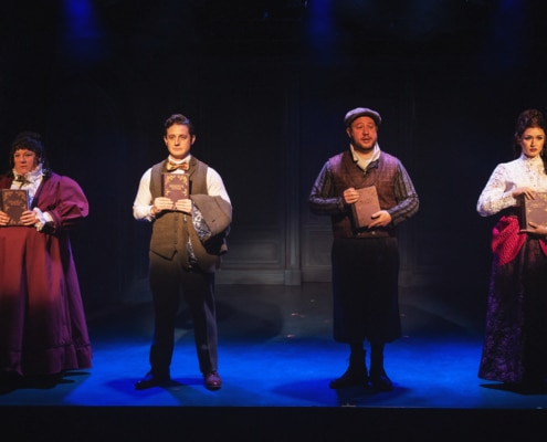 Four actors stand in a row on a dimly lit stage, each holding a book in front of their chest. They are dressed in Victorian-style costumes, including long skirts, vests, and high-collared blouses, against a dark backdrop with subtle paneling. The lighting creates a dramatic, moody atmosphere with blue and purple tones.