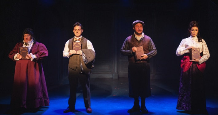 Four actors stand in a row on a dimly lit stage, each holding a book in front of their chest. They are dressed in Victorian-style costumes, including long skirts, vests, and high-collared blouses, against a dark backdrop with subtle paneling. The lighting creates a dramatic, moody atmosphere with blue and purple tones.