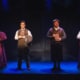 Four actors stand in a row on a dimly lit stage, each holding a book in front of their chest. They are dressed in Victorian-style costumes, including long skirts, vests, and high-collared blouses, against a dark backdrop with subtle paneling. The lighting creates a dramatic, moody atmosphere with blue and purple tones.