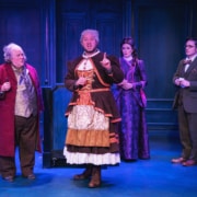 Four actors in Victorian-style costumes stand on a stage with a deep blue paneled backdrop. One actor in the center wears a layered dress with brown, orange, and cream tones and gestures with an index finger raised, while the others wear a mix of long coats, patterned vests, and elegant gowns. The scene is brightly lit, highlighting the ornate period clothing and theatrical setting.