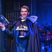 Actor in an ornate blue patterned suit and dark cape stands on stage holding a silver pedestal tray with a covered dessert and a glass dome lid. The background features paneled walls, a bookshelf, and a floral arrangement with red roses, all illuminated by dramatic blue lighting.