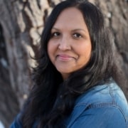 Person outdoors leaning against a tree trunk, wearing a denim shirt with long wavy hair cascading over the shoulders.